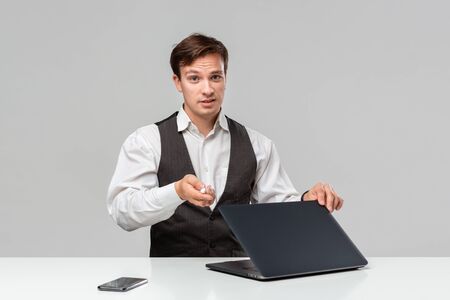 Businessman in a white t-shirt and grey vest working on a laptop. Man looking at the camera. Daily business routine.の写真素材