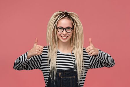 Beautiful young girl with dreadlocks in a striped shirt and jeans jumpsuit smiling holding thumbs up. Concept of likeの写真素材
