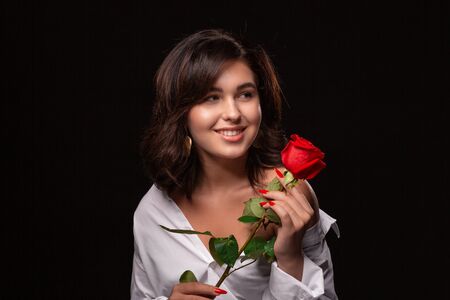 Beautiful woman with bright makeup posing isolated over black background with red rose. Image of young beautiful woman with red lips in a white shirtの写真素材
