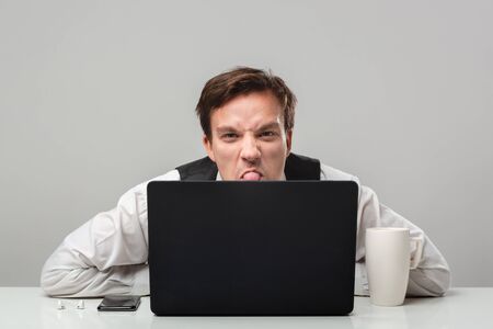 Businessman in a white t-shirt and grey vest is hiding behind laptop. Having problems. Daily...