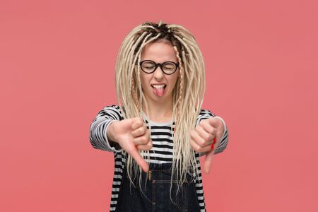 Beautiful young girl with dreadlocks in a striped shirt and jeans jumpsuit is upset holding thumbs down. Concept of dislikeの写真素材