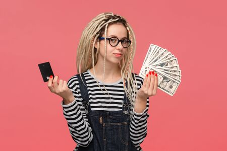 Teenager with dreadlocks in a striped shirt and jeans jumpsuit choosing between cash and bank card.の写真素材