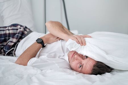 Young man in the white shirt lying on a bed with white linen and covers his head with a pollow. Concept of relaxationの写真素材