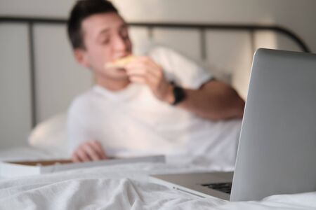 Young man in the white shirt and pajama smiling lying on a bed and eating tasty pizza. Focus on a...