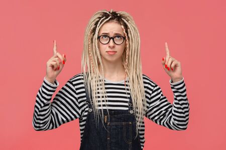 Beautiful young girl with dreadlocks in a striped shirt and jeans jumpsuit pointing with two fingers at copy space up. Place for advertisingの写真素材