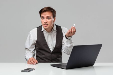 Businessman in a white t-shirt and grey vest takes off the headphones to hear better while working on the laptop. Time to work.の写真素材