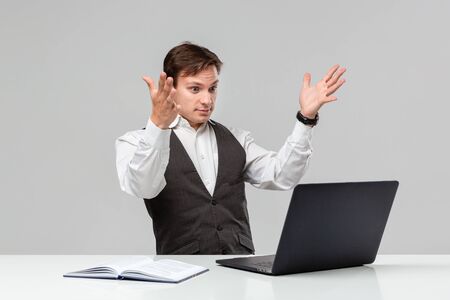 Businessman in a white t-shirt and grey vest is shocked checking information on a laptop sitting at the white table. Daily business routine.の写真素材