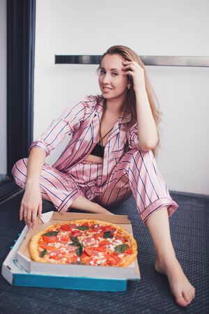 Young woman eating a piece of pizza sitting on floor wearing pajamas. Birthday party. Bachelorette party. Christmas party.の写真素材
