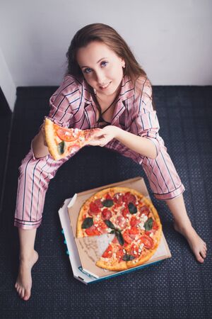 Young woman eating a piece of pizza sitting on floor wearing pajamas. Birthday party. Bachelorette party. Christmas party.の写真素材