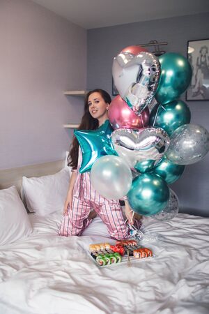 Birthday party. Beautiful young girl in pajamas on bed with balloons and sushi. Bachelorette party. Christmas party.の写真素材