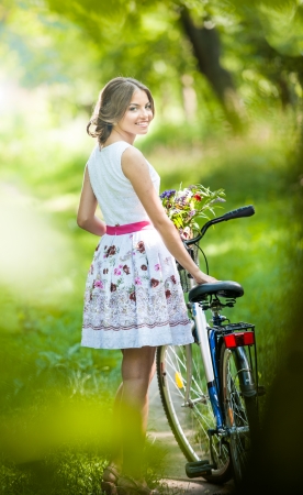 Beautiful girl wearing a nice white dress having fun in park with bicycle  Healthy outdoor lifestyle concept  Vintage scenery  Pretty blonde girl with retro look with bike and basket with flowersの写真素材