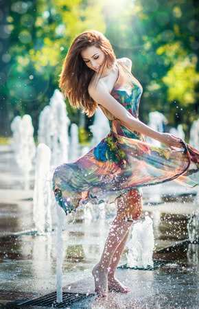 Attractive girl in multicolored short dress playing with water in a summer hottest day  Girl with wet dress enjoying fountains  Young beautiful happy female playing with outdoor water fountains の写真素材