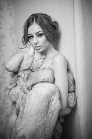 Attractive sexy young woman wearing a fur coat posing provocatively indoor. Portrait of sensual female with creative haircut, studio shot. Beautiful girl covered only with a fur exposing her shouldersの写真素材