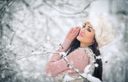 Woman with white fur cap smiling enjoying the winter scenery in forest. Side view of happy brunette...