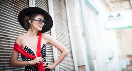 Attractive brunette girl with black hat, red scarf and sunglasses posing outdoor. Beautiful fashionable young woman with modern accessories, urban shot. Gorgeous brunette with large black hat smiling.の写真素材