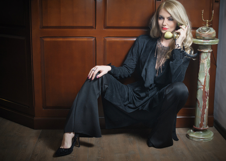 Young beautiful blonde woman in elegant black suit talking by phone sitting relaxed on the floor. Seductive fair hair girl leaning on wooden cabinet holding a vintage phone in classic interiorの写真素材