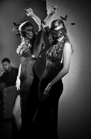 Gorgeous lady with long tight dress and mask in front of a large wall mirror, indoor shot. Mysterious young woman posing provocatively, black and white photo. Art photo of elegant sensual femaleの写真素材