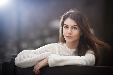 Attractive young Caucasian girl wearing a white blouse sitting on a bench in park. Beautiful brown hair teen girl with gorgeous eyes posing outdoor, art portrait, winter season. Pretty girlの写真素材