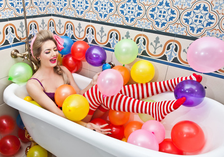 Blonde woman playing in her bath tube with bright colored balloons. Sensual girl with white and red striped stockings having fun in bathroom, covered with balloonsの写真素材