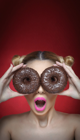 Beautiful model girl with bright pink lipstick holding two chocolate donuts. Funny joyful woman with sweets on dark red background.Young woman holding donuts against her eyes with surprised expressionの写真素材