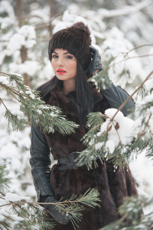 Beautiful woman in long black fur coat and cap enjoying the winter scenery in forest. Brunette girl posing under snow-covered trees branches in woods. Attractive young female in bright cold dayの写真素材