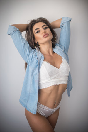 Attractive sexy brunette in white lingerie and denim shirt posing provocatively, near a grey wall,studio shot. Portrait of sensual woman with long hair, in classic boudoir scene.の写真素材