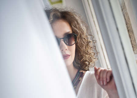 Slim young fashion model with curly hair wearing white blouse in a frame of window. Picture of lovely sexy fashionable woman with sunglasses and necklaceの写真素材