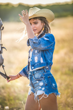 Beautiful blonde woman with curly hair with white hat and horse. Portrait of a girl with denim and her horse. Beautiful girl interacting and having fun with a horse at the ranchの写真素材