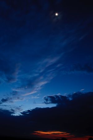  Moon shining through moving cloud cover against setting sun skyline with very  blue sky and dramatic cloudsの写真素材