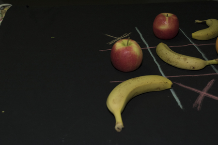 Tris Game with fruit. Three bananas, two apples and two oranges on a black background. Field lines...