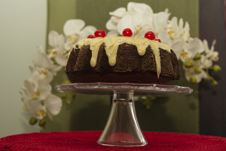 Close up of Bundt Cake with white chocolate and candied cherries on glass splashback and red crochet table centerpiece.の写真素材