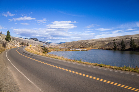 Tranquil Lake Between Long Road and Hills Under Light Blue Sky on one Sunny Day.の写真素材