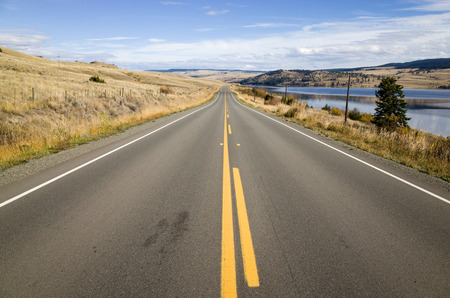Yellow markings on a tarred highway leading straight off into the distance alongside a lake in low hills in a scenic landscapeの写真素材