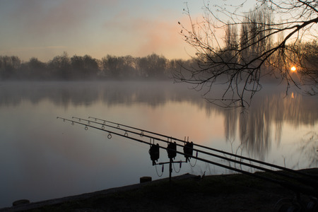 Sunrise over a lake with a silouhette of carp fishing rodsの写真素材