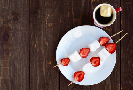 A cup of coffee and marshmallows with fresh strawberries on skewers on a wooden background.の写真素材