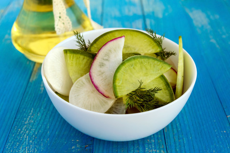 Salad of radish of different varieties (daikon, green, Chinese red) chopped slices, flavored with sunflower oil in a white bowl on a blue wooden background. Close-up.の写真素材