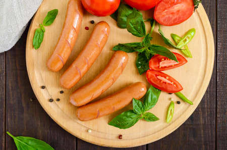 Juicy sausages with tomatoes and basil on a round cutting board on a dark wooden background. Top view.の写真素材