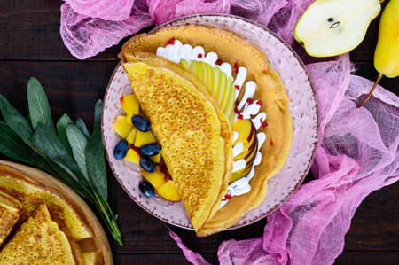 Golden crepes with pieces of peach, pear, cream and grapes on a ceramic plate on a dark background. Top view.の写真素材