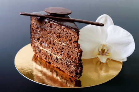 A piece of chocolate cake with chocolate cream on a golden napkin with a white orchid flower on a black background.の写真素材