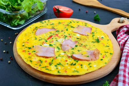 Omelet with ham, cheese, greens on a wooden board on a black background.  Fritata - a delicious, healthy breakfast.の写真素材