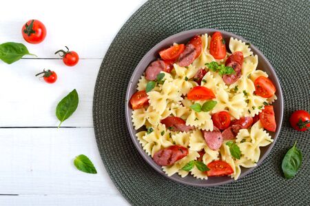 Tasty pasta farfalle with grilled sausages, fresh cherry tomatoes and basil on a plate on a white background. Top view, flat lay.の写真素材