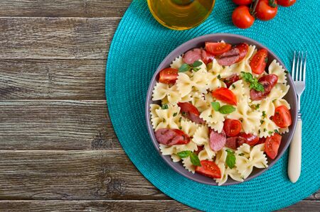 Tasty pasta farfalle with grilled sausages, fresh cherry tomatoes and basil on a plate on a wooden background. Top view, flat lay.の写真素材