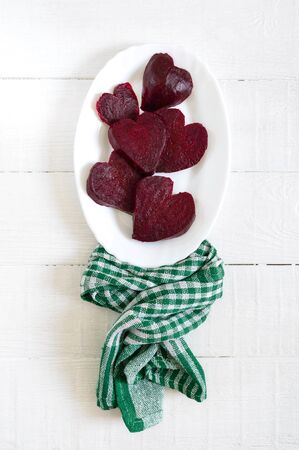 Slices of cooked beets in the shape of a heart on a white plate and a green kitchen towel on a white wooden background.の写真素材