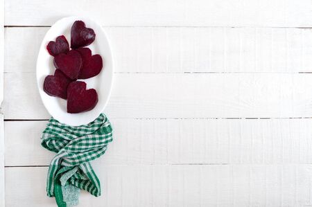 Slices of cooked beets in the shape of a heart on a white plate and a green kitchen towel on a white wooden background.の写真素材