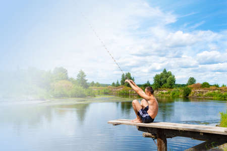 Boy teenager sits on a wooden bridge going to jump into the river from the swinging rope on sunny summer day. Copy space.の写真素材