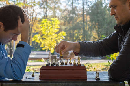 Two adult man play chess outdoors in the park. Side view.