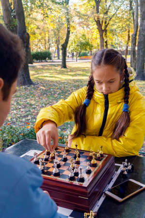 A young guy and a beautiful girl in a yellow jacket with pigtails play chess outdoors in an autumn park. The concept of competition between brother and sister, friends. Vertical view.の写真素材