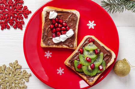 Creative festive sandwiches in the shape of a Christmas tree and Santa hat, made with fresh fruits and marshmallows on a red plate on a white wooden background. Children's breakfasts concept.の写真素材