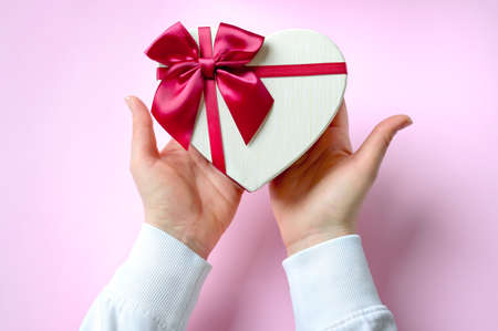 Womans hands holding gift or present heart shaped box decorated big red bow on pink table top view....