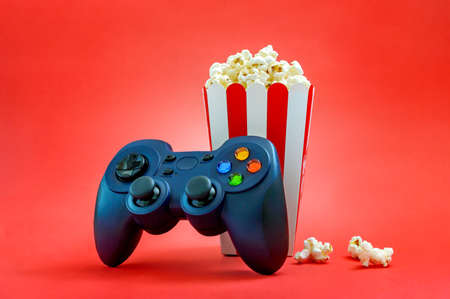Gamepad and popcorn in a striped paper cup on a red background. Gaming, leisure and entertainment...